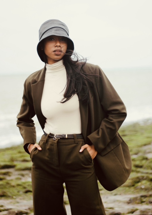 Model in olijfgroen pak met bucket hat aan de kust